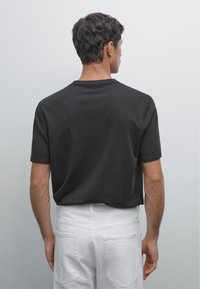 Uomo con capelli corti e scuri, che indossa una camicia nera a maniche corte e pantaloni bianchi, in piedi di fronte a uno sfondo grigio chiaro e uniforme.