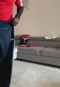 Person in a red shirt and dark pants standing near a gray sofa with folded clothes and sports jerseys on it.