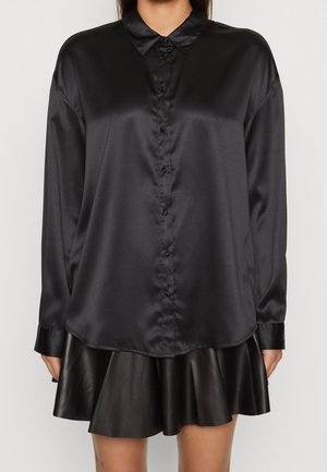 Frau trägt ein schwarzes Satinhemd mit langen Ärmeln und Knöpfen sowie einen schwarzen, plissierten Minirock aus Leder, stehend vor einem einfarbigen Hintergrund.