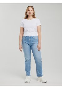 Pikkade juustega naine, kes kannab valget t-särki, helesiniseid teksapükse ja valgeid tenniseid, seisab ühevärvilise helehalli tausta ees.