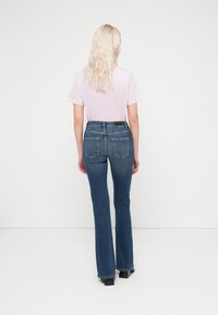 Μπλε τζιν με φαρδιά καμπάνα, μέτριο λούσιμο, σχεδιασμός με πέντε τσέπες και ψηλή μέση. Συνδυασμένα με ανοιχτό ροζ μπλουζάκι, σε όψη από πί σω.