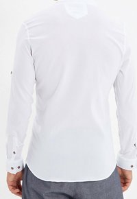 Biała koszula z długim rękawem wykonana z teksturowanej tkaniny, z mankietami na guziki i ostrym kołnierzem. Posiada przeszycie na tylnej karczku.