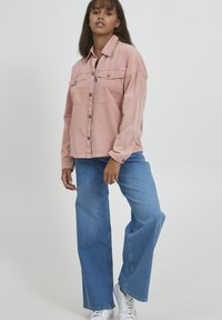 Lichtroze overhemdd met twee borstzakken, gecombineerd met wijde blauwe jeans en witte sneakers, met een ontspannen snit en lange mouwen.