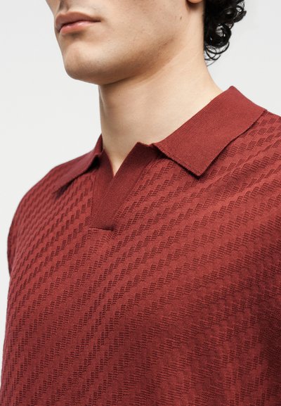 Άνδρας που φοράει πλεκτό μπλουζάκι πόλο σκούρου κόκκινου χρώματος με υφή και κοντό γιακά, εμφανιζόμενος από τους ώμους μέχρι το πηγούνι, μπροστά σε απλό φόντο.
