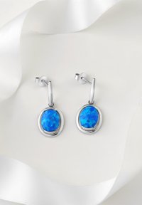 Silverörhängen med ovala blå opaler omgivna av en slät silvrig infattning, visade mot en ljus bakgrund med vitt satinband.