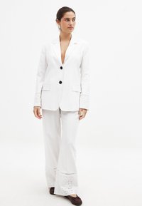 Balta pritaikyta švarkas su dviem juodais mygtukais, išpjautomis atlapomis ir akytomis detalėmis ant rankovių, derinamas su atitinkamomis platėjančiomis kelnėmis.