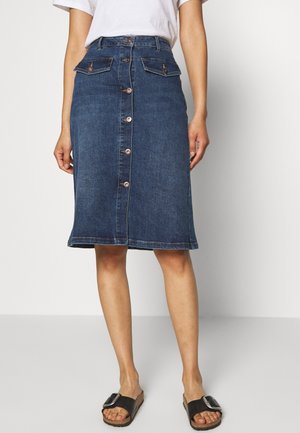 Vrouw draagt een knielange spijkerrok met knopen aan de voorkant en klepzakken, gecombineerd met zwarte sandalen met gesp, staand tegen een witte achtergrond.