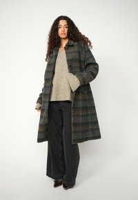 Wollplaidmantel in Grau und Grün mit Kragen, Knopfverschlüssen und großen Taschen, über einem beigen Pullover und weiten schwarzen Hosen.