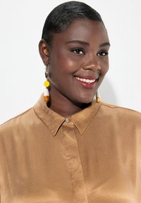 Camicia button-up marrone e setosa con colletto, caratterizzata da un piccolo bottone frontale e una texture liscia. Grandi orecchini rotondi gialli e bianchi.