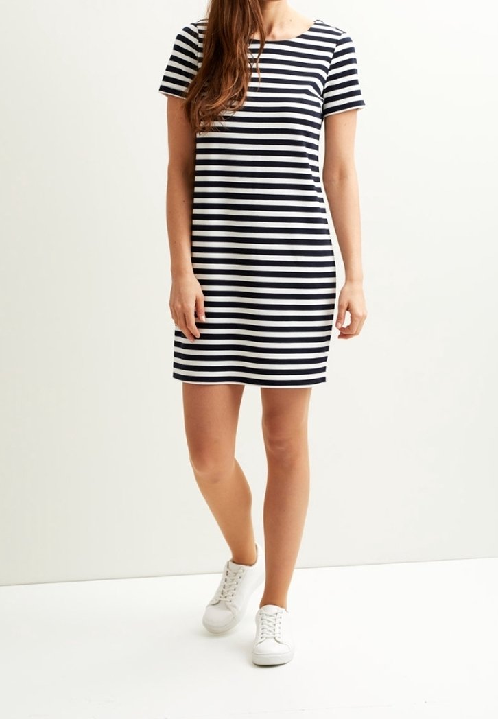 Femme portant une robe à manches courtes à rayures horizontales noir et blanc et des baskets blanches, debout devant un fond clair uni.