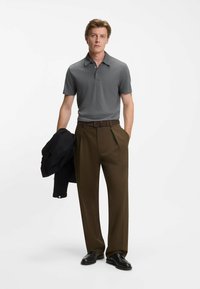 Graues Poloshirt mit Kragen, kombiniert mit dunkelbraunen Hosen und schwarzen Schuhen. Eine schwarze Jacke ist über einen Arm gelegt.