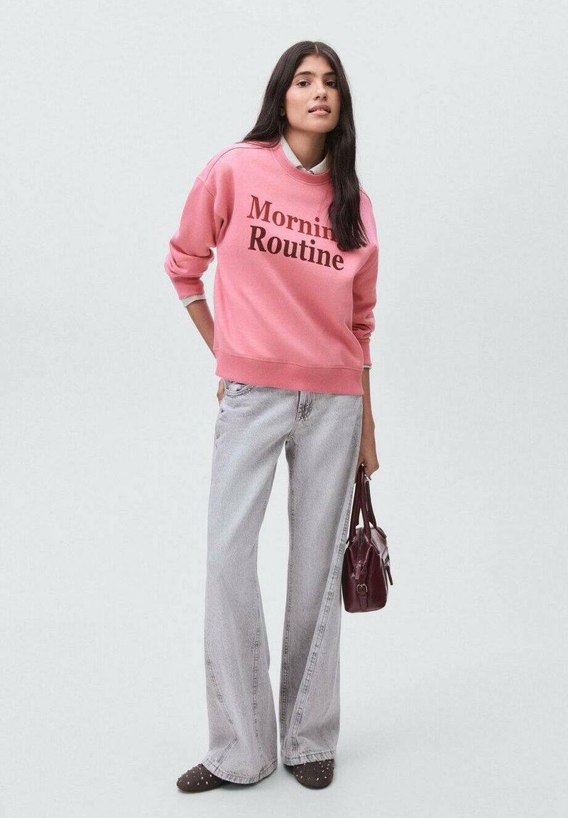 Mladá žena v růžovém svetru "Ranní rutina", světle šedých kalhotách s širokými nohavicemi, tmavých botách se cvočky a držící tmavě červenou kabelku.