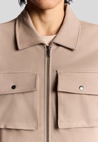 Beige jacka med dragkedja, två framfickor och en mjuk textur. Har en pekig krage och svarta tryckknappar på fickorna.