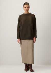 Brūns ribots adījums ar apaļu kakla izgriezumu, apvienots ar beigu maksi svārkiem un brūniem zābakiem ar papēdi. Modelis stāv pret neitrālu fonu.