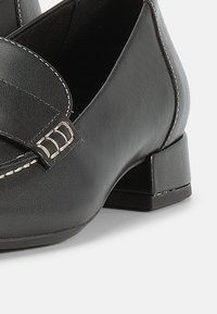 Μαύρα δερμάτινα μοκασίνια με χαμηλό τετράγωνο τακούνι, διακοσμημένα με λουράκι και αγκράφα. Οι ραφές προσθέτουν υφή στην επιφάνεια.