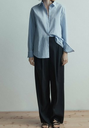 Blue and white striped button-up shirt, untucked, with long sleeves; paired with black wide-leg trousers and flat sandals.