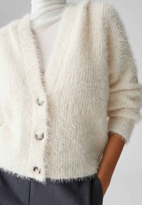 Weicher, cremefarbener Cardigan mit V-Ausschnitt, drei strukturierten Knöpfen, weitem Ärmel und lässiger Passform. Weiches, gestricktes Material.