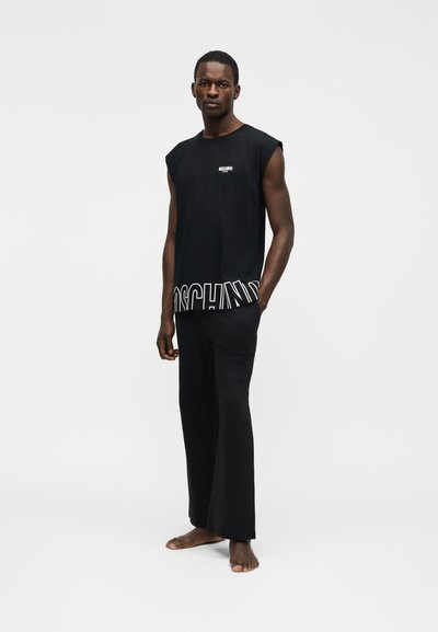 Άνδρας που στέκεται ξυπόλητος φορώντας μαύρο αμάνικο τοπ με έντονο κείμενο και φαρδιά μαύρα παντελόνια, σε απλό φωτεινό φόντο.