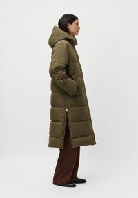Woman wearing olive green long quilted coat with hood, dark brown wide-leg pants, and black shoes, standing in profile with hands in pockets.