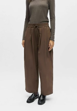 Person som bærer brune bukser med vid ben og snor i livet, en tettsittende taupefarget langermet topp, svarte loafers og mønstrede svarte sokker foran en ensfarget bakgrunn.