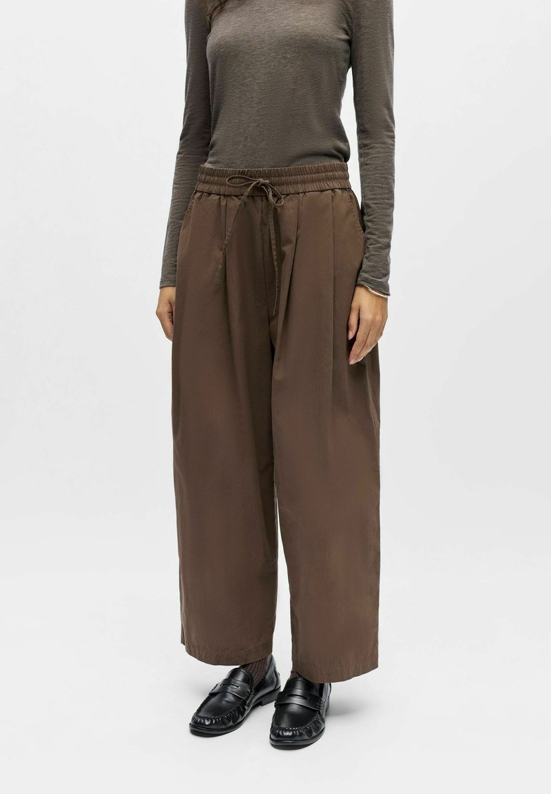 Personne portant un pantalon marron à jambes larges avec cordon, un haut taupe ajusté à manches longues, des mocassins noirs et des chaussettes noires à motifs sur un fond uni.