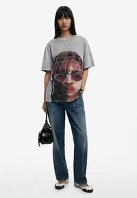 Sivé nadrozmerné tričko s grafickým potlačou tváre, v kombinácii s modrými džínsami a čiernymi doplnkami, vrátane malej kabelky.