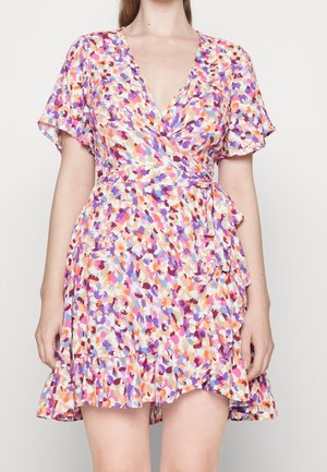 Kobieta ubrana w krótką, kolorową sukienkę w kwiaty z zakładanym przodem, falbanowym dołem i rozkloszowanymi rękawami na jednolitym tle.