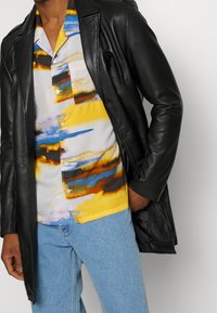 Svart läderjacka över en kortärmad skjorta med ett flermönstrat abstrakt mönster i gult, blått och brunt, kombinerat med ljusblå jeans.