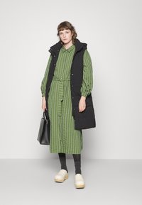 Vestido camisola verde aos riscas com cintura cingida, combinado com um colete acolchoado preto. Os acessórios incluem uma bolsa preta e tamancos bege.