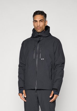 Man wearing black weatherproof jacket with hood, front zipper, and multiple zippered pockets, standing against a plain light background.