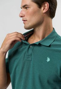 Tyrkysová polokošeľa vyrobená z jemnej tkaniny, s dvojitým uzáverom a malým vyšitým logom na ľavej hrudi. Golier je mierne zvýšený.