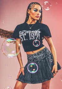 Jeune femme aux cheveux tressés portant un crop top noir Ed Hardy et une jupe en jean plissée, entourée de bulles de savon flottantes sur fond rose.