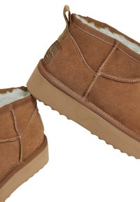Botas de tobillo de ante marrón con un forro suave y peludo, suela de goma texturizada y un parche de marca en el lateral. Diseño curvado con costuras simples.