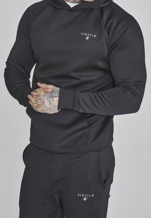 Mand med tatoverede hænder iført sort SikSilk hættetrøje og matchende sorte bukser med logo på bryst og lår, stående mod grå baggrund.