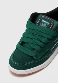 Baskets en daim vert avec de larges lacets verts, un patch logo Reebok noir et vert, des coutures blanches et une semelle en caoutchouc marron clair.