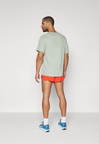 Kortermet lysegrønn t-skjorte med avslappet passform, kombinert med lysende oransje shorts. Modellen har på seg blå løpetreningssko og hvite sokker.