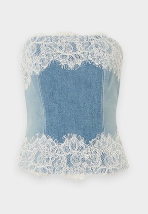 Kobieta w niebieskim gorsetie jeansowym bez ramiączek z białą koronkową lamówką oraz białych, szerokich spodniach z wysokim stanem, stoi z rękami w kieszeniach.