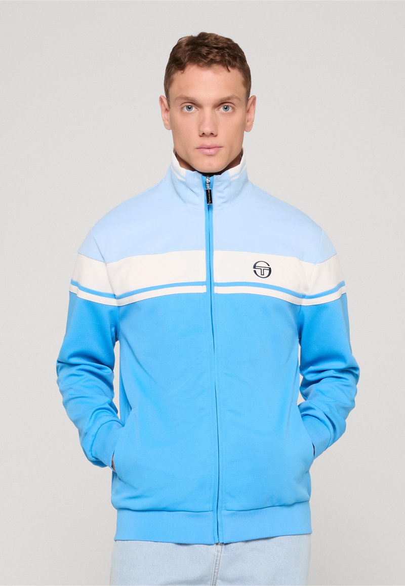 Lys blå glidelåsjakke med hvite horisontale striper over brystet og en liten logo på venstre side. Mykt, glatt stoff.