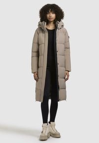 Lang beige pufferjakke med hætte, der har knaplukning og quiltet design. Båret over sorte leggings og kraftige beige støvler.