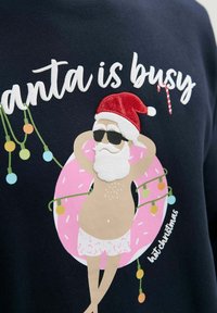 Marineblauwe sweatshirt met een illustratie van de kerstman in een rode hoed en zonnebril, die op een roze donut ligt, met de tekst "Kerstman is druk".