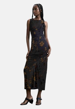 Donna con lunghe trecce che indossa un vestito nero senza maniche con motivi floreali dorati e tacchi neri aperti, in piedi davanti a uno sfondo semplice.
