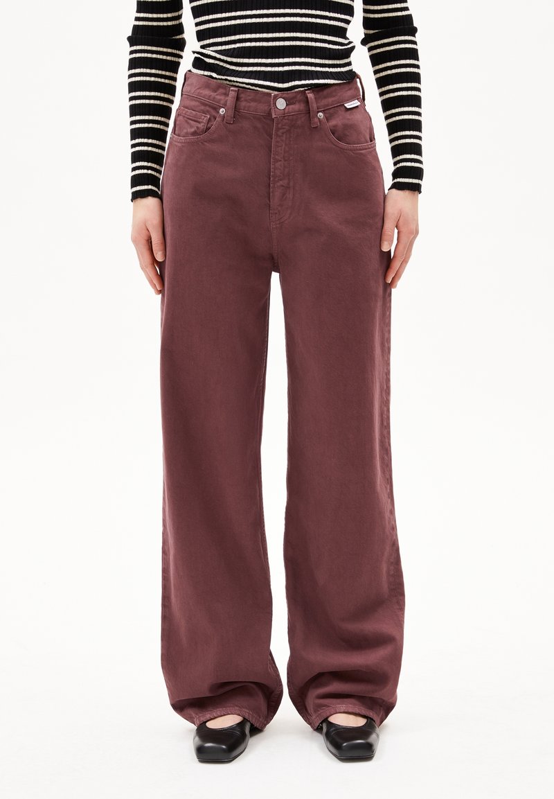 Personne portant un pantalon bordeaux taille haute à jambes larges, des chaussures noires à bout carré et un haut à manches longues rayé noir et blanc, debout droit.