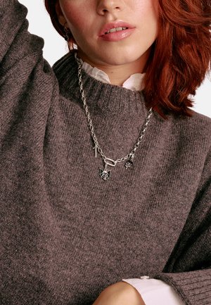 Massive Halskette in Silberoptik mit Anhängern, darunter eine Fledermaus, Waffen und ein Spinnenmotiv, präsentiert über einem strukturierten braunen Pullover.