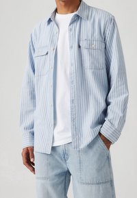 Camicia a righe blu chiaro con bottoni e due taschini sul petto, caratterizzata da una vestibilità rilassata e tessuto morbido, indossata sopra una maglietta bianca.