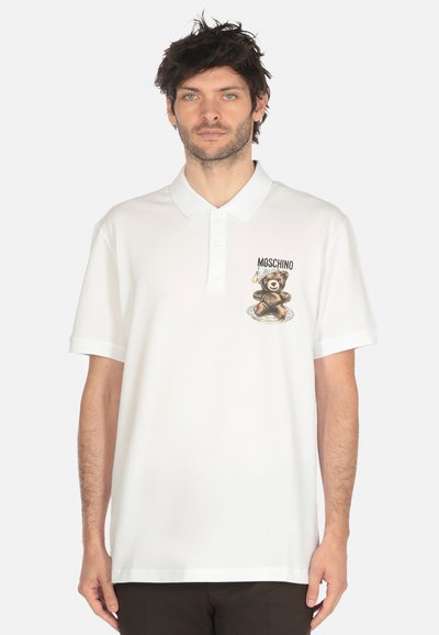 Uomo che indossa una polo bianca con un orsacchiotto e il testo "MOSCHINO", in piedi contro uno sfondo bianco uniforme.