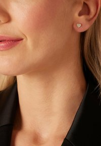 Silberne, herzförmige Steckerohrringe mit einem hellblauen Emaille-Mittelteil und kleinen klaren Akzenten, getragen am Ohr einer Person mit glatter Haut und hellem Haar.