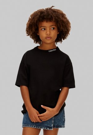 Dziecko z kręconymi włosami ubrane w czarny t-shirt Calvin Klein i niebieską dżinsową spódniczkę, stojące ze splecionymi rękami i patrzące w prawo.
