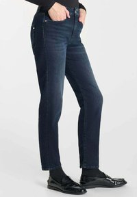 Jean en denim bleu foncé avec une coupe slim, design traditionnel à cinq poches et détails de couture subtils. Associé à des mocassins noirs et des chaussettes.