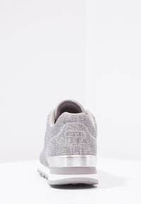 Grå atletisk sko med ett texturerat tygöverdrag, silverdetaljer på hälen och en vit sula. Rund form, minimalistisk design.