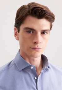 Camicetta azzurra con un motivo testurizzato, colletto con bottoni e bottoni bianchi. Progettata per un aspetto aderente, che mette in risalto un look curato.